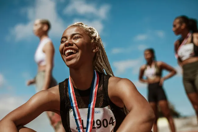 Smiling female athlete with medal around neck celebrating victory with fellow runners in background.