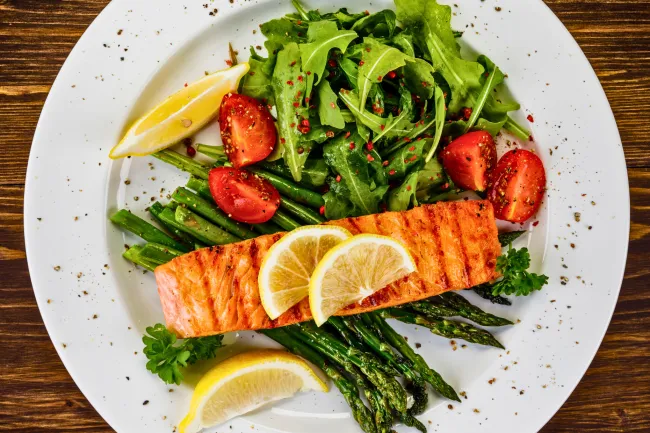 Grilled salmon served with lemon slices, asparagus, cherry tomatoes, and arugula salad on a white plate.
