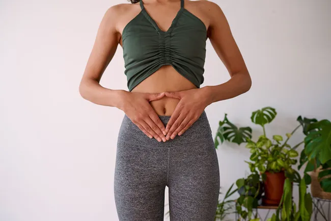 Woman in green crop top and gray leggings holding hands over stomach with houseplants in background
