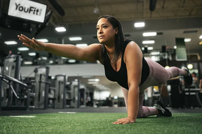 Woman performing bird dog exercise on turf at a modern gym with YouFit sign in the background
