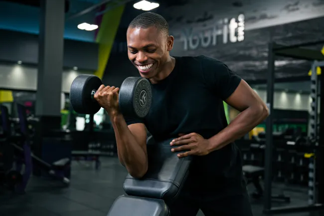 Smiling young man lifting a dumbbell using a preacher curl bench in a modern gym setting.