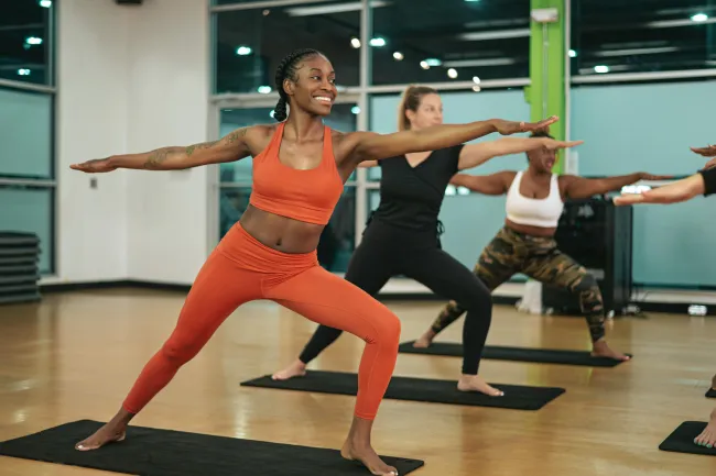 Group of diverse women practicing yoga warrior pose in bright fitness studio with mats on wooden floor
