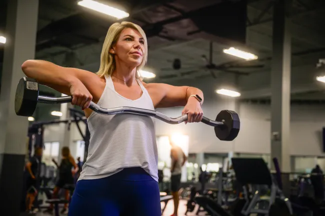 Woman in white tank top lifting barbell with weights in gym, focusing on strength training.