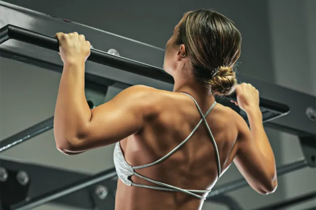 Woman performing pull-up exercise showcasing toned back muscles in gym setting with workout equipment.