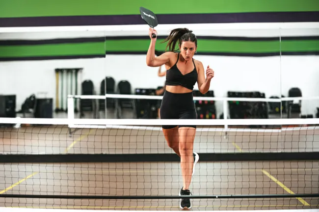 Athletic woman in black sportswear lunges to hit a pickleball on an indoor court.