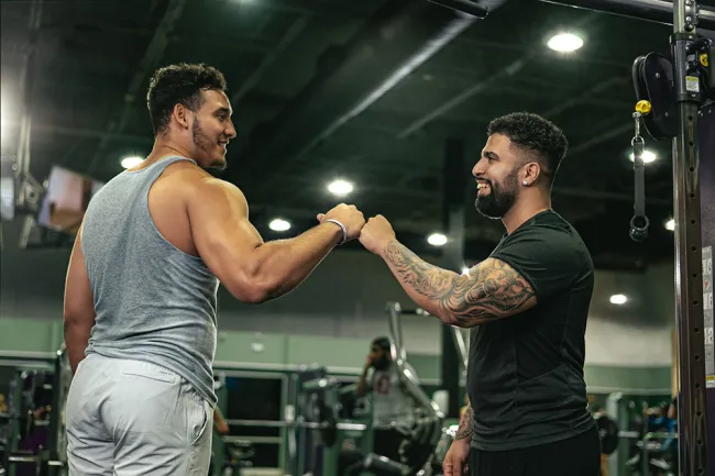 Two muscular men fist bump in a gym setting, celebrating a workout success with equipment in the background.