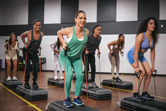 Group of diverse women exercising with resistance bands on step platforms in a fitness class studio.
