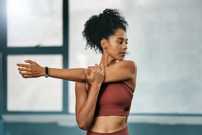 Woman in rust sportswear stretching her arm indoors with natural light from window.