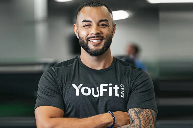 Smiling man with beard wearing a YouFit Gyms shirt and a tattooed arm in a gym setting.