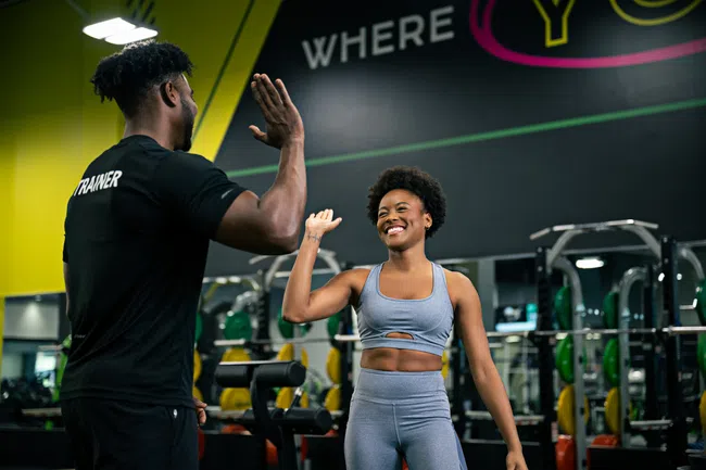 Female athlete and male trainer exchanging a high-five in a colorful gym setting with fitness equipment.
