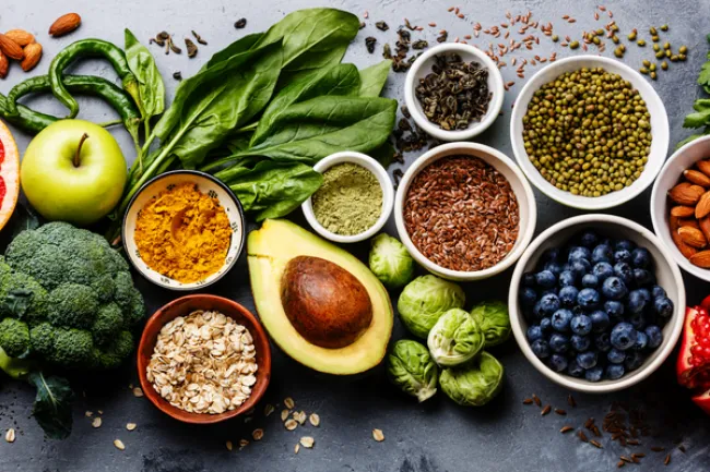 Fresh healthy superfoods including avocado, spinach, blueberries, broccoli, oats, nuts, and spices on a gray surface.