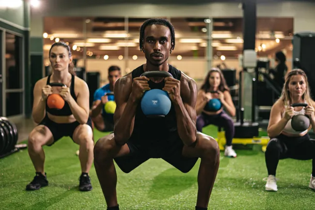 Group of diverse people doing kettlebell squats together in a gym with green turf flooring