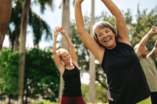 Smiling mature woman stretching arms up during outdoor group fitness class with palm trees in background