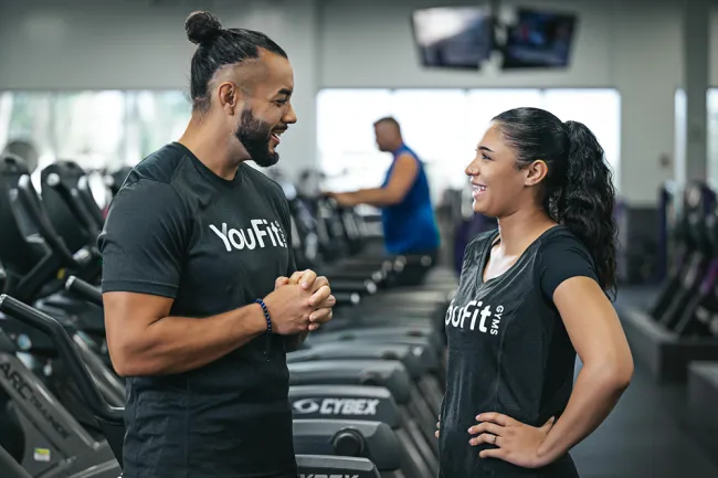 Male and female trainers wearing YouFit shirts talk and smile by gym treadmills in a fitness center.
