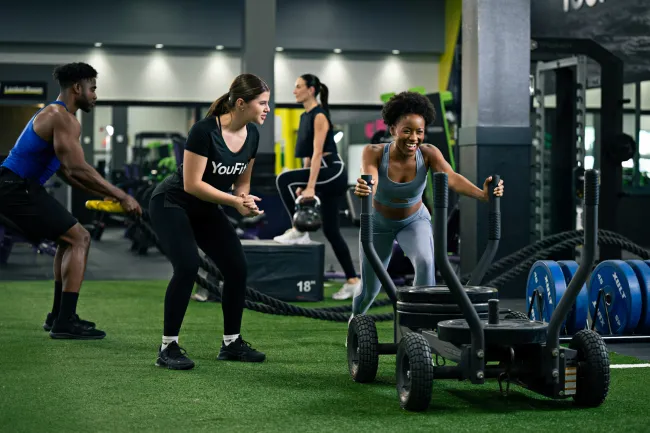 Group of diverse people exercising with battle ropes, sled push, and kettlebells in modern gym setting.