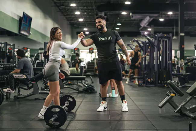 Two gym-goers fist bump in a busy fitness gym with weights and exercise equipment around.