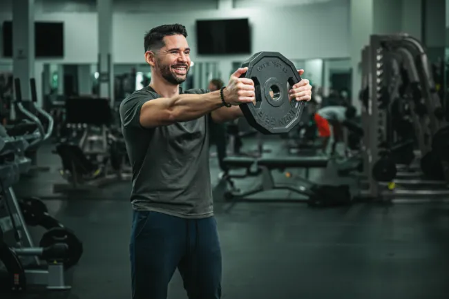 Smiling man lifting a weight plate in a modern gym with various exercise equipment in the background
