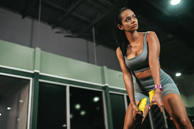 Fit woman in gray workout clothes using battle ropes in a gym with large windows and high ceilings