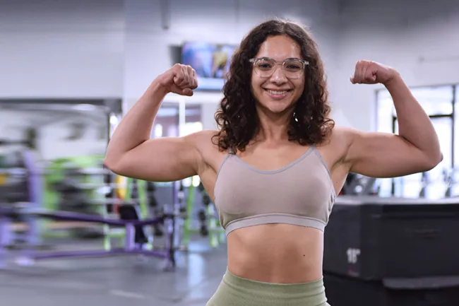 Smiling woman in gym flexing biceps wearing glasses and workout clothes showing fitness strength.