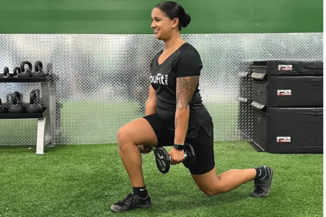 Woman performing a weighted lunge exercise holding dumbbells in a gym with green walls and artificial turf.