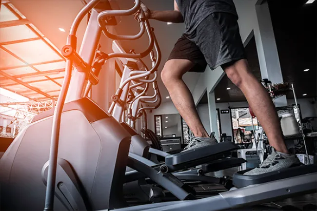Person exercising on elliptical machine in gym with sunlight streaming through large windows.