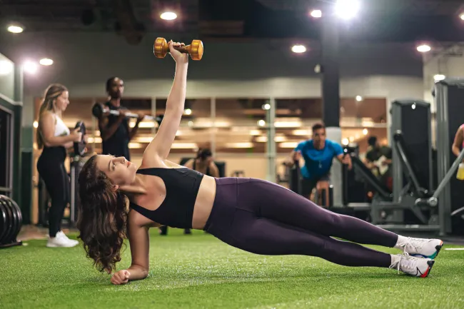 Woman performing side plank with a dumbbell in gym, other people exercising in background