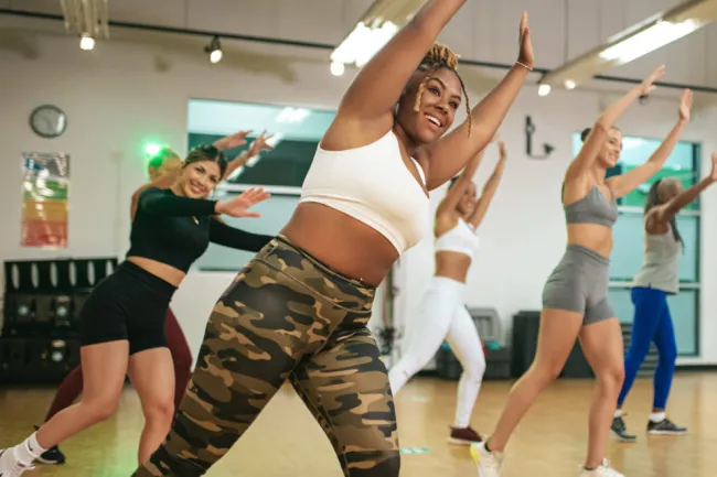 Diverse group of women in activewear smiling and stretching arms up during a fitness class in a studio.