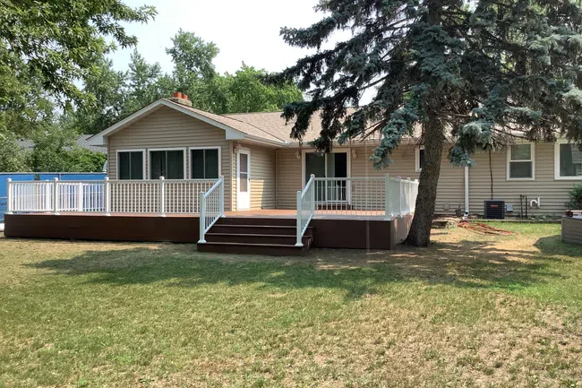 Spacious backyard with deck and railing attached to a single-story beige house with large windows and trees.