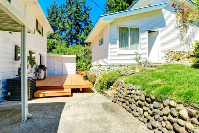 Sunny backyard with white houses, stone retaining wall, wooden deck, and green lawn under clear blue sky