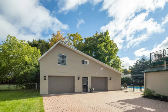 Modern beige three-car garage with paved driveway surrounded by green trees under a blue cloudy sky