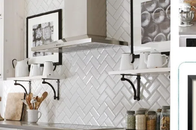 White kitchen with herringbone tile backsplash, stainless steel hood, open shelves with white ceramics, and glass jars.
