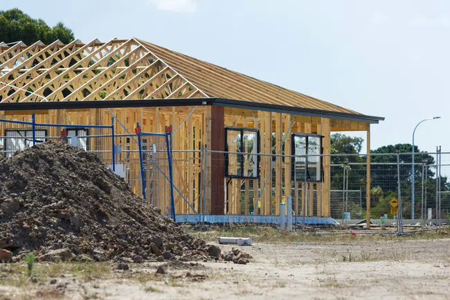 Wooden frame structure of a house under construction on a sunny day with a dirt pile in the foreground.