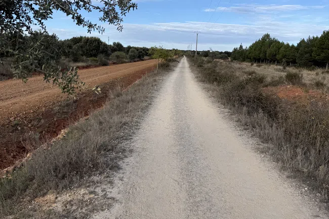 Long straight dirt road flanked by dry grass and trees under a blue sky with scattered clouds.