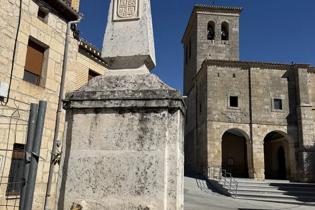 Historic stone fountain with rooster statue and coat of arms near old church under blue sky.