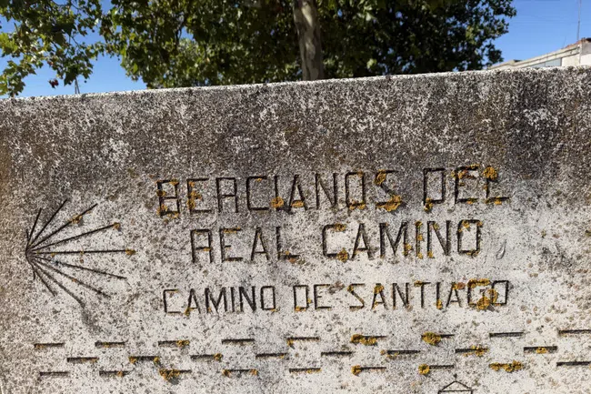 Stone plaque with weathered text marking the Real Camino de Santiago pilgrims route under trees.