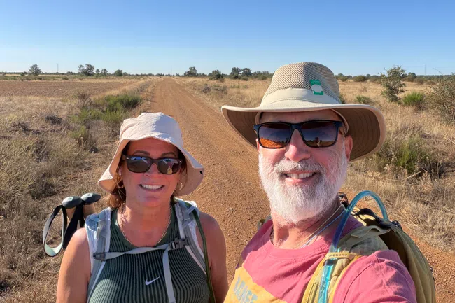 Smiling hikers wearing hats and backpacks on a dry dirt trail under a clear blue sky in a rural landscape
