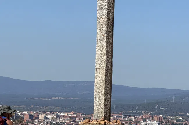 Stone cross monument with small stones on its base overlooking a cityscape under clear blue sky.