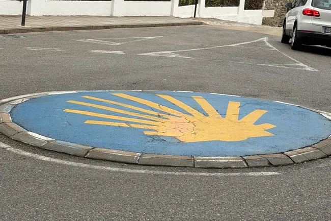 Roundabout with a yellow scallop shell symbol on a blue background in a suburban street with a white house and car nearby.