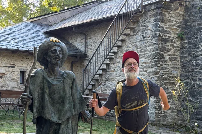 Man in hiking gear posing with bronze statue of pilgrim holding walking stick outside stone building with stairs.