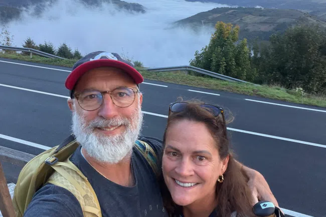 Smiling couple selfie with backpacks on mountain road overlooking fog-filled valley and cloudy sky.