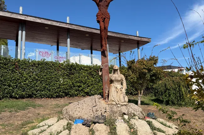 Rusty metal crucifix with Jesus and stone sculpture of kneeling woman on a rock under a clear blue sky