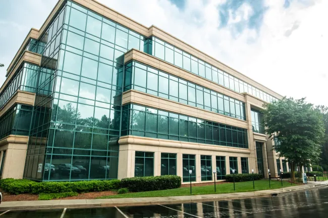 Modern office building with large glass windows and beige stone accents reflected on a wet street on a cloudy day