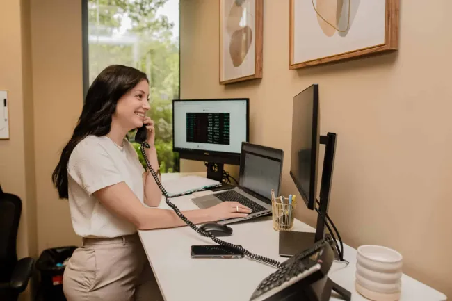 Woman at home office desk using phone with triple monitors showing financial data, smiling and working