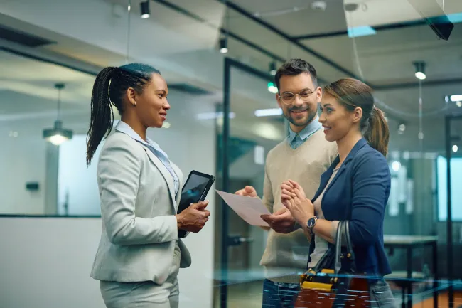 Three professionals engage in a discussion inside a modern office with papers and a tablet.