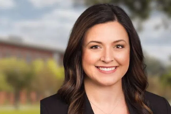 Professional woman smiling with a modern office building, trees, and water fountains in the background under a cloudy sky.