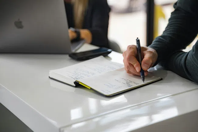 Person writing notes in a notebook at a white desk with a laptop and smartphone in the background.