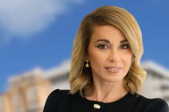 Confident businesswoman stands with folded arms in front of blurred city buildings under a blue sky.