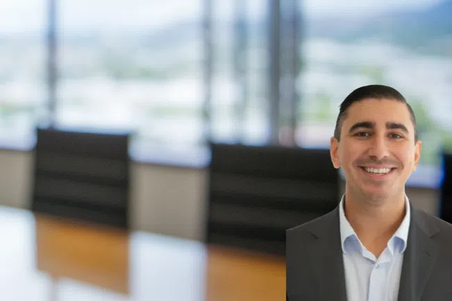 Smiling man in business suit with blurred office conference room and large windows in background.