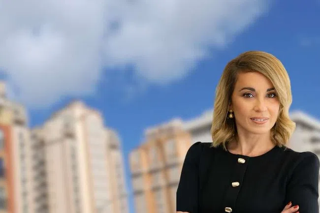 Confident businesswoman stands with folded arms in front of blurred city buildings under a blue sky.