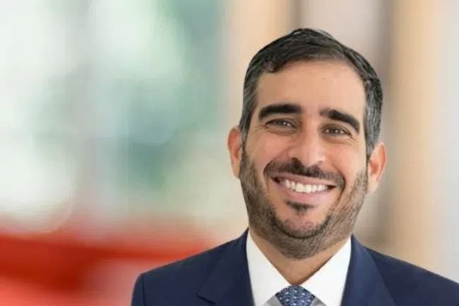 Smiling professional man in suit standing in a bright office lobby with orange seating and large windows.